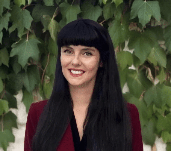 Woman with long black hair and bangs wearing a burgundy blazer stands in front of leafy green vines.